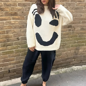 Oversized white knit sweater featuring a large black smiley face emoji design on the front, worn by a woman with apricot skin tone and visible shoulder length hairstyle. The sweater has long sleeves and a relaxed fit in 2XL.