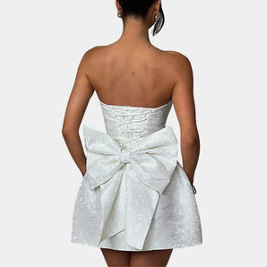 Strapless white satin cocktail dress featuring a delicate lace overlay and a large bow, elegantly worn by a woman with styled hair.