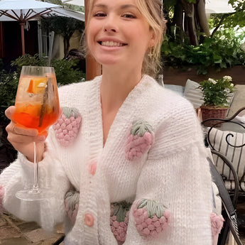 
A woman wearing a pink XL strawberry knit cardigan with a trendy V-neck design, smiling while standing near a table adorned with plants and flowers.