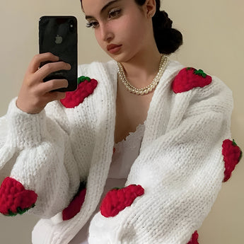 A woman wearing a strawberry knit cardigan with a trendy V-neck design, featuring long sleeves. Her hands are visible, and she is surrounded by white flowers and plants, which add a natural touch to the image.