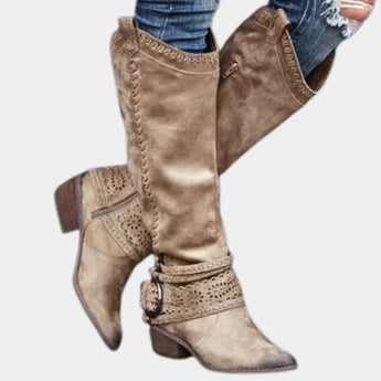 Taupe suede knee-high boots with a western-inspired design, featuring a studded heel and side zipper, displayed in a brown color and size 42. The boots are paired with trousers, emphasizing their stylish and fashionable appeal.