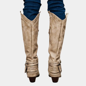 Taupe suede knee-high boots with a Western-inspired design, featuring a studded heel and a side zipper, shown on a human leg.