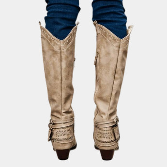 Taupe suede knee-high boots with a Western-inspired design, featuring a studded heel and a side zipper, shown on a human leg.