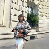 Textured bomber jacket in a stylish monochrome design, featuring a combination of black and brown patterns. Suitable for winter street style, the jacket is shown in a large size, with full sleeves and a snug fit around the waist. The model pairs the jacket with casual pieces like a T-shirt and denim, accessorizing with eyewear and carrying a bag.