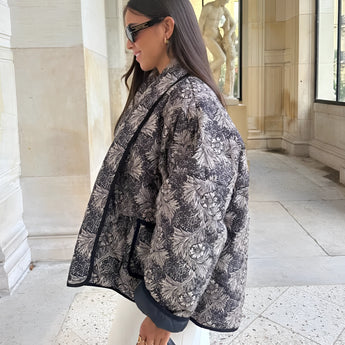 Textured black patterned bomber jacket in large size, featuring a stylish monochrome winter street style. The jacket has a collar, long sleeves, and is worn by a person with eyewear standing next to a window.