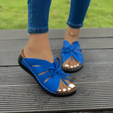 A pair of blue women's flat sandals featuring a bow-tie design, worn by a person standing on a surface, with part of the legs and a dress visible.