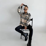 Oversized plaid jacket modeled by a woman, featuring long sleeves and a relaxed fit. The model is sitting on a chair, wearing eyewear, embodying a chic autumn streetwear style.
