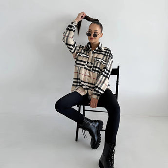 Oversized plaid jacket modeled by a woman, featuring long sleeves and a relaxed fit. The model is sitting on a chair, wearing eyewear, embodying a chic autumn streetwear style.