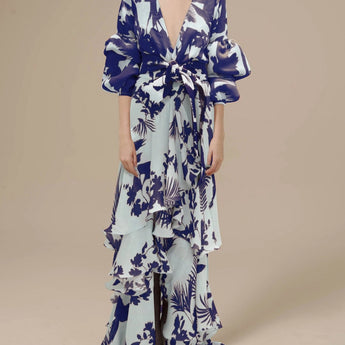 Tropical floral maxi dress with sleeves in blue, featuring a wrap design, modeled by a person with long hair. The dress is a one-piece garment with a defined waist and a stylish collar, suitable for daytime fashion.