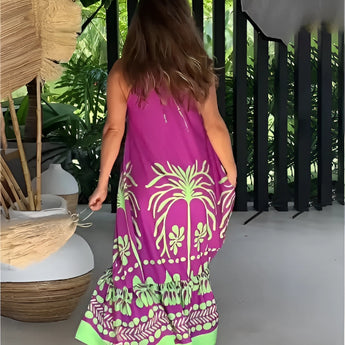 Tropical maxi dress with a vibrant purple and green palm design, featuring loose sleeves and a flowing silhouette. The dress is being worn outdoors near a fence, surrounded by greenery and grass, showcasing a colorful pattern with tints and shades of magenta.