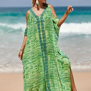 A woman wearing a tropical print green beach kaftan stands near the water, smiling under a clear blue sky, showcasing the lightweight V-neck summer dress with loose sleeves and a relaxed waist.