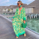 Tropical print green and white long sleeve kaftan modeled on a sunny day, showcasing luxurious beachwear with fashion-forward design. The kaftan has a relaxed waistline, and the model is accessorized with sunglasses, set against a backdrop of clear sky and scattered clouds.