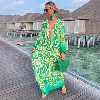 Tropical print green and white long sleeve kaftan modeled on a sunny day, showcasing luxurious beachwear with fashion-forward design. The kaftan has a relaxed waistline, and the model is accessorized with sunglasses, set against a backdrop of clear sky and scattered clouds.