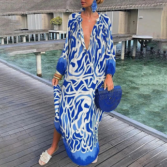 
A woman wearing a tropical print green and white long sleeve kaftan stands near a body of water. She is accessorized with a sun hat and sunglasses. The blue and azure tones of the kaftan complement her beachwear ensemble. The kaftan is a 2XL size.