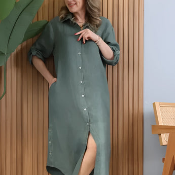 Turquoise linen dress with asymmetrical hem, featuring long sleeves, worn by a model standing. The dress is styled for summer, highlighting the shoulder and waist.