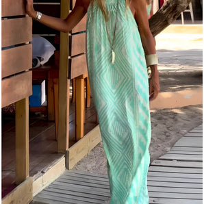 
A woman wearing a turquoise tie-dye maxi dress, which is lightweight and sleeveless, suitable for beachwear. The dress features a flowing design, with details like exposed shoulders and a cinched waist. She is accessorized with eyewear and stands on a wooden surface. The color and pattern of the dress evoke a cool, breezy vibe.