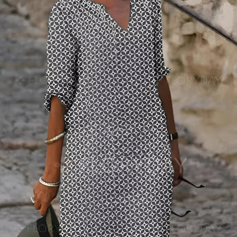 A woman wearing a green and white V-neck geometric print loose fit summer dress with 3/4 sleeves, standing and looking sideways, with her hand on her hip.