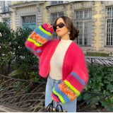 A person wearing a vibrant multicolor patchwork boho chic granny square wool cardigan in red, standing indoors near a window with plants, accessorized with glasses.
