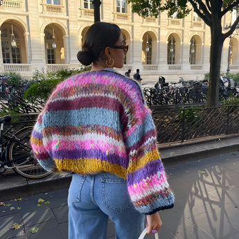 Handmade wool boho chic sweater in vibrant multicolor patchwork, featuring long sleeves and a purple color, size large, displayed outdoors near a building with plants and trees in the background.