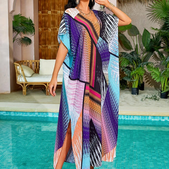 Woman wearing a vibrant print kaftan, standing near water and surrounded by tropical plants. The dress features purple and aqua colors, designed for luxury resort and tropical beach wear, showcasing stylish shoulder and sleeve details.