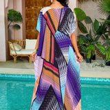 Woman wearing a vibrant print kaftan dress with purple and tropical patterns, standing near a window with houseplants in the background. The dress features a loose, flowing design with a focus on leisure and resort fashion.
