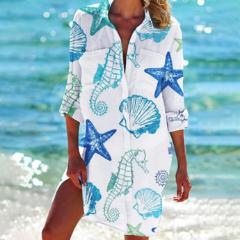 Woman wearing a vibrant tie-dye beach cover-up dress in blue seahorse pattern, standing on the beach with water in the background, smiling and enjoying the summer.
