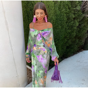 Vibrant watercolor sheer off-shoulder maxi evening dress in shades of purple and pink, featuring a flowing one-piece design with sleeves, modeled by a smiling woman outdoors amidst greenery.