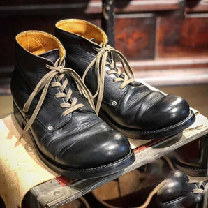 A pair of vintage black leather men's high-top distressed lace-up boots shown close-up, worn with blue jeans. The boots have a rugged and worn look, suitable for outdoor walking and casual athletic styling.