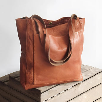 Vintage brown distressed leather tote bag with shoulder straps, handcrafted luxury design, displayed on a wooden surface.