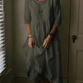 Woman wearing a dark green, loose-fitting vintage linen dress with short sleeves, standing near a window with blinds. The dress features a bohemian style, perfect for summer, and is in a size 5XL.