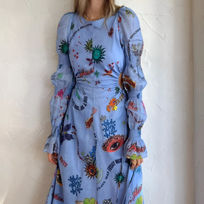 
A woman smiling while wearing a whimsical blue maxi dress featuring colorful embroidery and patterns, standing outdoors with flowers and plants in the background.
