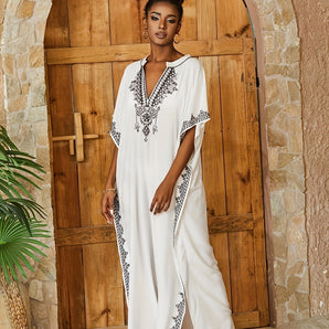 A woman wearing a white embroidered bohemian kaftan with a V-neck, featuring detailed Moroccan designs, standing near houseplants.