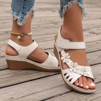 White floral wedge sandals featuring a beaded strap and elegant cutout design, shown on a model's foot and leg, highlighting the shoe's beige color and stylish appearance.