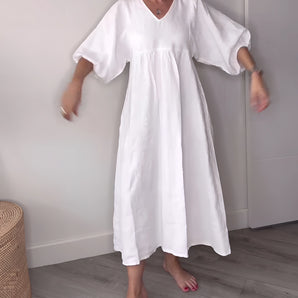 White linen V-neck dress with mid-length hem, featuring short sleeves and a flattering waistline. The dress is worn by a smiling person, showcasing the dress's casual summer style.