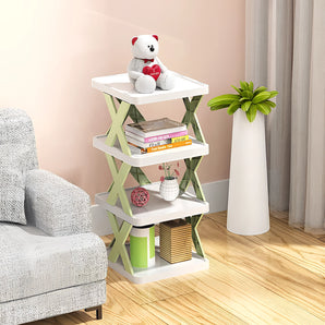 White X-shaped corner bookshelf with a modern minimalist design, featuring wooden shelves and a variety of items such as houseplants, a flowerpot, and toys displayed on it. The bookshelf is situated on wooden flooring.