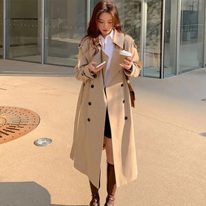 Women's beige double-breasted long trench coat in an XL size, featuring a chic design suitable for autumn wear. The coat has a classic collar, long sleeves, and a defined waistline, styled with a dress shirt underneath, reflecting a fashionable and sophisticated look.