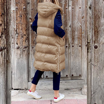 
A woman wearing a beige hooded puffer vest, which is a stylish sleeveless winter coat. The vest is complemented by other casual winter wear. The background includes a wooden door, adding a warm, rustic touch to the scene.