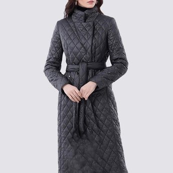 
A woman wearing a long beige quilted coat with a belted waist, featuring a slim trench design. The coat has a classic collar and long sleeves, and is paired with sunglasses and a hat. The model is positioned to highlight the coat's elegant silhouette and insulated detailing.