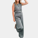 A woman wearing a gray sleeveless jumpsuit designed for casual summer wear, featuring a flattering fit with a defined waist and knee-length pants. She is smiling and posing with her arms bent, highlighting the jumpsuit's comfortable and stylish design suitable for warm weather.