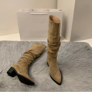 
A pair of women's beige suede knee-high boots with a slouchy design and block heels, displayed on a wooden floor.