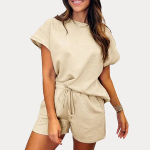 Women's beige summer romper set featuring a textured fabric and drawstring waist, worn by a smiling model with styled hair. The romper has short sleeves and showcases the collar and thigh-length design.