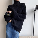 
A woman wearing a black chunky knit oversized turtleneck sweater paired with jeans, standing outdoors with sunglasses on.