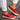 Women's red suede slip-on sneakers with perforated design, shown on a foot standing on a white surface.