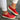 A pair of women's blue suede slip-on shoes featuring a colorful band with shades of red, orange, yellow, and pink, a perforated design, and a white sole, shown on a person's feet while standing.