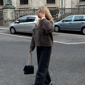 A woman wearing a brown corduroy oversized jacket stands casually in an urban setting, with a parked car and buildings in the background.