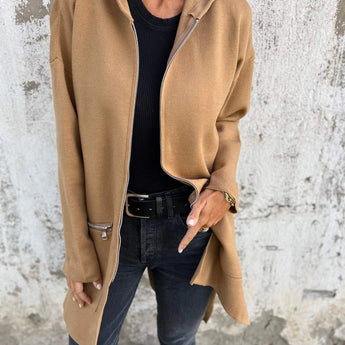 
A woman wearing a lightweight wool camel coat, styled as a casual chic outerwear piece, paired with jeans. The coat features long sleeves and extends to the waist. The model is accessorized with sunglasses and a neutral-toned outfit underneath the coat.