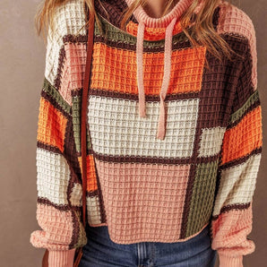 
A woman wearing a cozy high neck patchwork sweater with a multicolor geometric design, featuring shades of orange. The sweater has long sleeves and is paired with jeans.