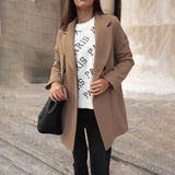 A woman wearing a chic urban style double-breasted long red winter coat, standing confidently with her hand in her waist pocket. She accessorizes the outfit with sunglasses and a beige dress shirt underneath, suitable for a fashionable city look.