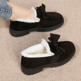 A pair of women's black faux fur-lined suede loafers featuring a moccasin style with a decorative bow tie, displayed against a natural wood background.