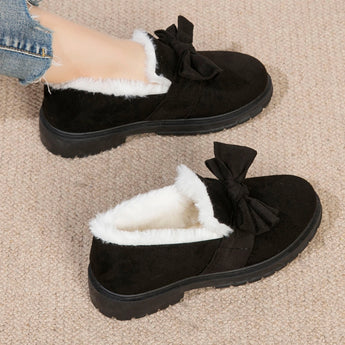 A pair of women's black faux fur-lined suede loafers featuring a moccasin style with a decorative bow tie, displayed against a natural wood background.